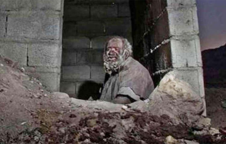 সাজপোশাক বা খাবারদাবারের মতোই হাজির থাকার জায়গাও কম অভিনব নয়। যেন কবরখানায় ঢুকছেন, এমন গর্তে থাকেন তিনি। বসবাসের আরও একটা জায়গা রয়েছে তার। বৃদ্ধের থাকার জন্য ইটের সারি দিয়ে খোলা ছাউনি গড়ে দিয়েছেন গ্রামবাসীরা। ছবি: সংগৃহীত