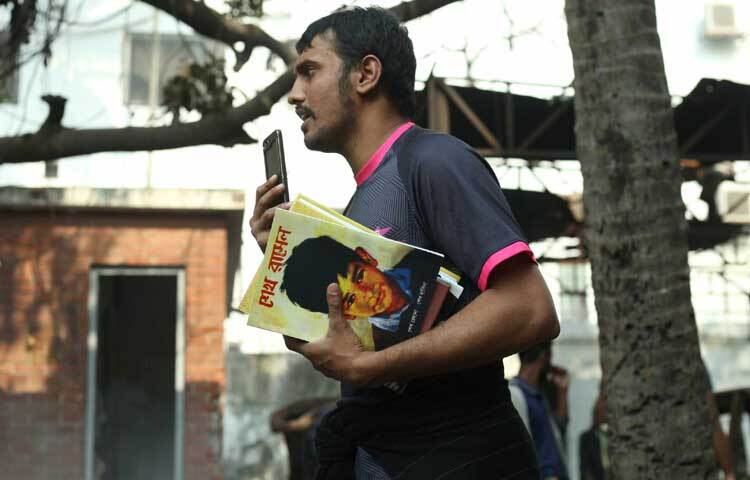 কিছু বই হাতে দাঁড়িয়ে আছে এক যুবক।