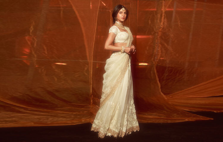 প্রিয়াঙ্কার পরা অফ-হোয়াইট শাড়িটির সরলতার ভেতর রয়েছে রাজকীয় গাম্ভীর্য। লেসের সূক্ষ্ম কাজ আর সোনালি সুতা বোনা বর্ডার একে দিয়েছে সমৃদ্ধতা, যেন পুরনো দিনের অভিজাত রুচি আর আধুনিক নান্দনিকতার নিখুঁত মেলবন্ধন।