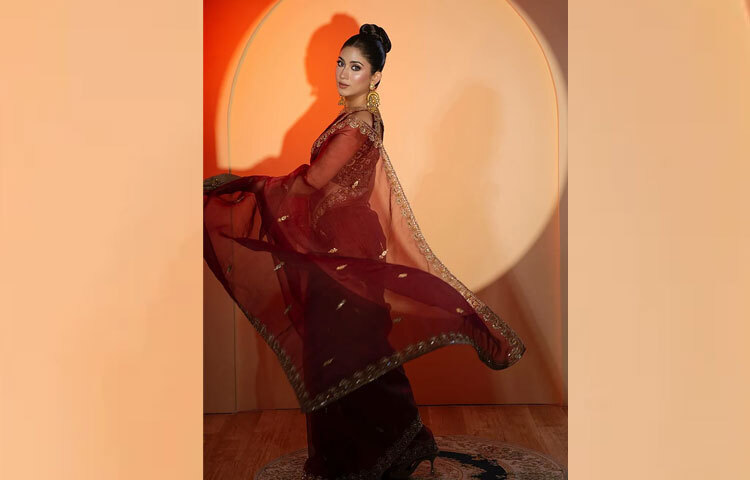 চুল বাঁধা হয়েছে স্টাইলিশ স্লিক বানে, যা পুরো লুকের ভারসাম্য বজায় রেখেছে। কানে দুলছে সোনালি ঝোলানো দুল, হাতে রইল সূক্ষ্ম চুড়ি ও আংটির ছোঁয়া-এই ছোট ছোট ডিটেইলসেই সম্পূর্ণ হয়েছে তার আভিজাত্যের গল্প।