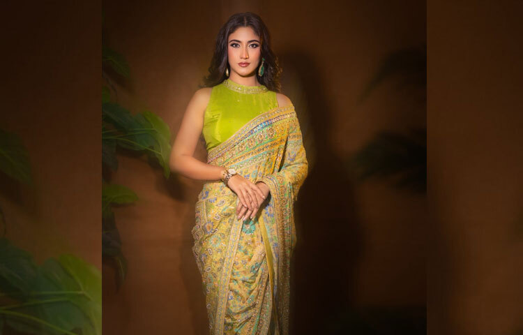 ফ্লোরাল প্রিন্টের হালকা অলিভ বেসে সূক্ষ্ম হাতে করা সোনালি, ফিরোজা, সবুজ ও কপার টোনের এমব্রয়ডারি। তার ওপর ছড়িয়ে থাকা সিকুইনের ঝলক যেন সকালের সূর্যালোকের মতো কোমল অথচ দীপ্ত। শাড়িটির প্রতিটি ভাঁজেই যেন মিশে আছে সৌন্দর্যের পরিশীলিত নান্দনিকতা।