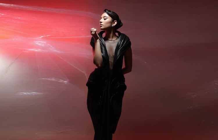 গাউনের মাঝখানের স্বচ্ছ অংশটি সাহসী, কিন্তু ভঙ্গি নয়। এটি কেবল ত্বক দেখানোর আয়োজন নয়; এটি নিয়ন্ত্রণের প্রদর্শনী। কাঁধের কাঠামোগত ভলিউম মুখের দিকে দৃষ্টি টানে, আর নীচের দিকে নামা মসৃণ ভাঁজ চলনে যোগ করে নাটকীয়তা।